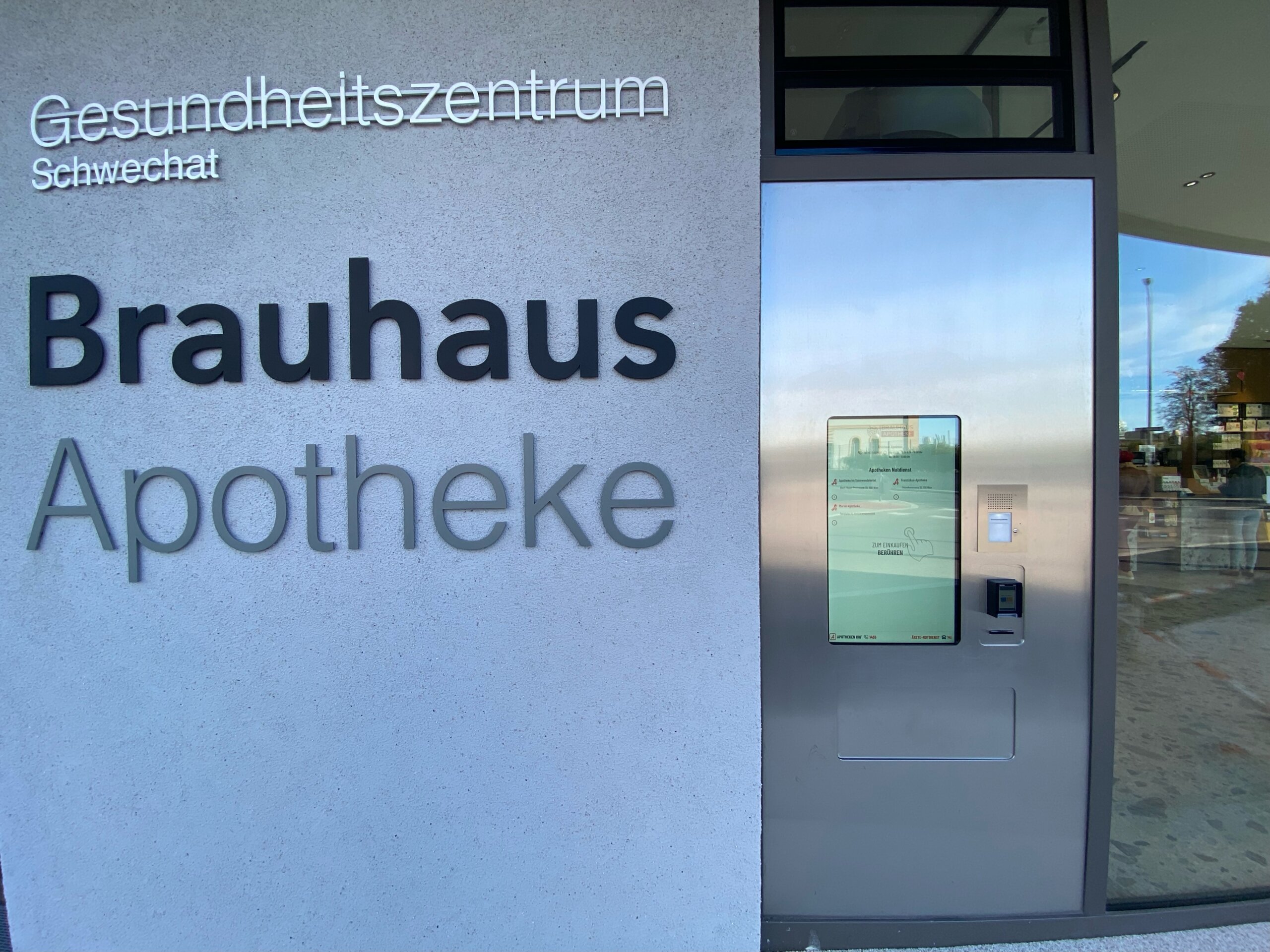 Die Außenmauer der Apotheke, auf dieser "Gesundheitszentrum Schwechat" und "Brauhaus Apotheke" steht, rechts daneben der Außenautomat